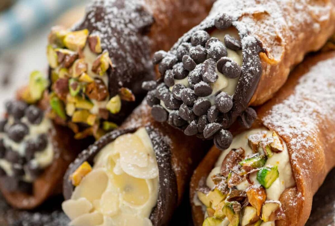 Golden crispy cannoli shells filled with creamy white ricotta mixture, dusted with powdered sugar and garnished with chopped pistachios, arranged on a rustic wooden serving platter
