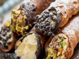 Golden crispy cannoli shells filled with creamy white ricotta mixture, dusted with powdered sugar and garnished with chopped pistachios, arranged on a rustic wooden serving platter
