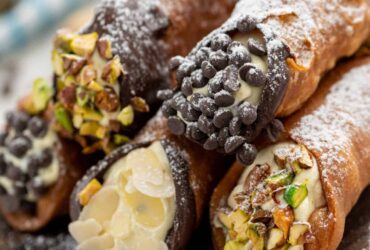 Golden crispy cannoli shells filled with creamy white ricotta mixture, dusted with powdered sugar and garnished with chopped pistachios, arranged on a rustic wooden serving platter