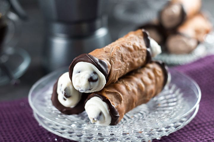 Close-up shot of a single cannoli cut in half showing the creamy ricotta filling studded with chocolate chips and pistachios, with powdered sugar dusting the plate