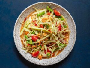 Overhead shot of Thai papaya salad som tam in a white bowl, garnished with crushed peanuts, halved cherry tomatoes, lime wedges, and fresh Thai basil on a wooden board