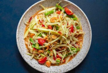 Overhead shot of Thai papaya salad som tam in a white bowl, garnished with crushed peanuts, halved cherry tomatoes, lime wedges, and fresh Thai basil on a wooden board