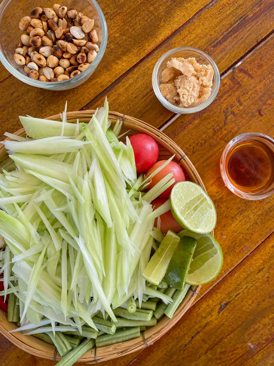 Flat lay of ingredients for Thai papaya salad including shredded green papaya, bird's eye chilies, lime halves, fish sauce bottle, cherry tomatoes, and crushed peanuts on a light surface