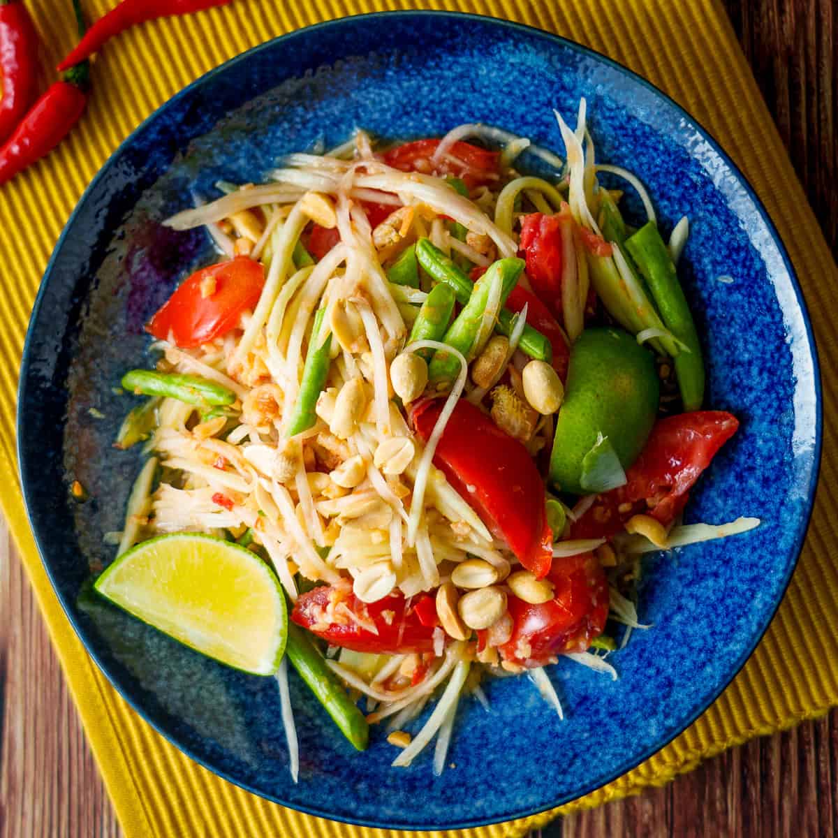 Close-up of Thai papaya salad som tam served in a small wooden bowl with a pair of chopsticks, garnished with fresh cilantro, peanuts, and a wedge of lime on a textured linen napkin