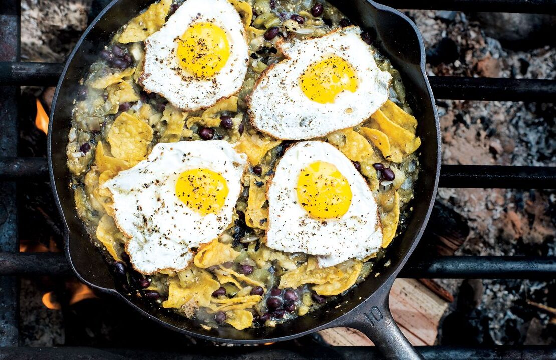Campfire chilaquiles in a cast iron skillet with runny eggs, red salsa, crumbled cotija cheese, sliced jalapeños, and fresh cilantro, photographed over a rustic wooden camp table with morning light