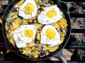 Campfire chilaquiles in a cast iron skillet with runny eggs, red salsa, crumbled cotija cheese, sliced jalapeños, and fresh cilantro, photographed over a rustic wooden camp table with morning light