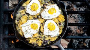 Campfire chilaquiles in a cast iron skillet with runny eggs, red salsa, crumbled cotija cheese, sliced jalapeños, and fresh cilantro, photographed over a rustic wooden camp table with morning light