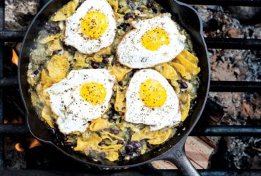 Campfire chilaquiles in a cast iron skillet with runny eggs, red salsa, crumbled cotija cheese, sliced jalapeños, and fresh cilantro, photographed over a rustic wooden camp table with morning light