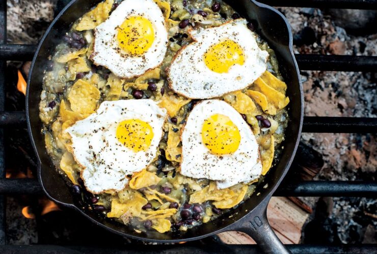 Campfire chilaquiles in a cast iron skillet with runny eggs, red salsa, crumbled cotija cheese, sliced jalapeños, and fresh cilantro, photographed over a rustic wooden camp table with morning light