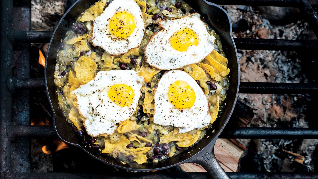 Campfire chilaquiles in a cast iron skillet with runny eggs, red salsa, crumbled cotija cheese, sliced jalapeños, and fresh cilantro, photographed over a rustic wooden camp table with morning light