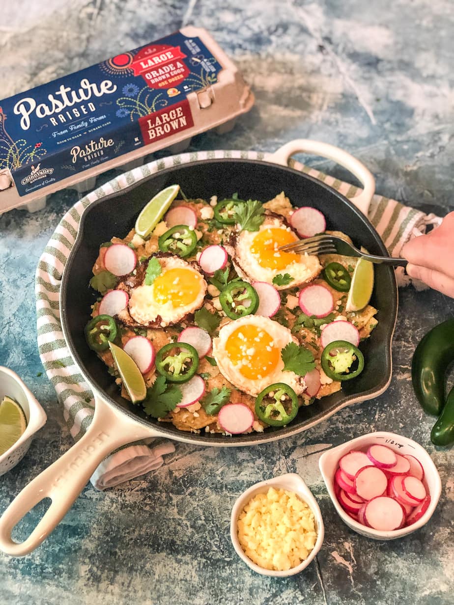 Overhead flat-lay of campfire chilaquiles ingredients including tortilla chips, jar of red salsa, six eggs in a carton, cotija cheese, canned black beans, sliced jalapeños, fresh cilantro, and a cast iron skillet on a rustic wooden surface