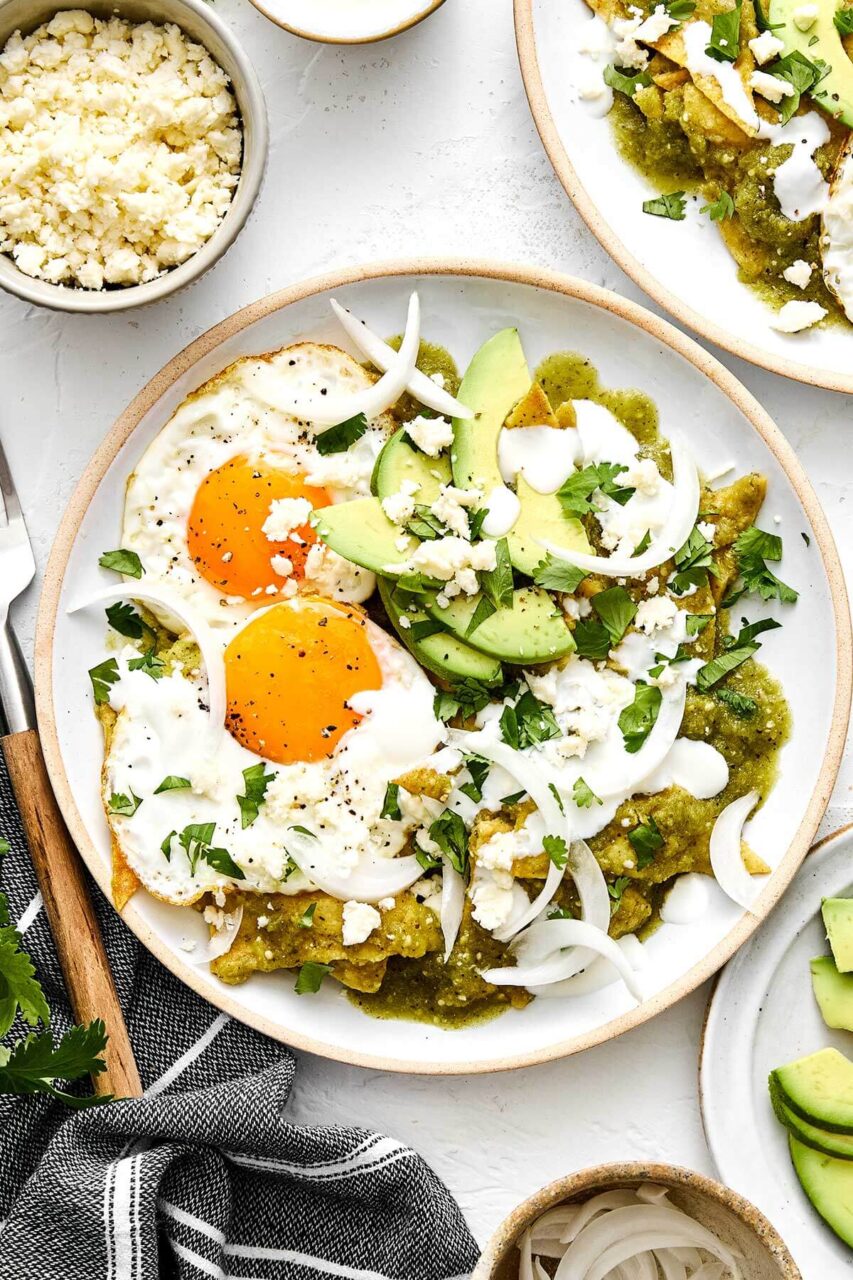 Close-up of a single serving of campfire chilaquiles in a bowl with a fork breaking a perfectly runny egg yolk over crispy tortilla chips, topped with crumbled cotija, sliced avocado, and fresh cilantro