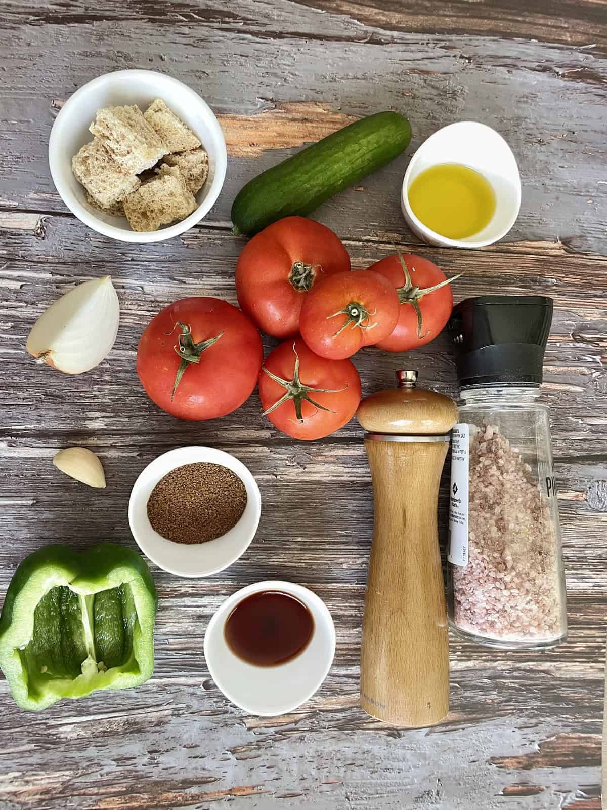 Overhead flat lay of fresh gazpacho ingredients including ripe tomatoes, cucumber, red bell pepper, garlic cloves, olive oil bottle, and red wine vinegar on a marble surface
