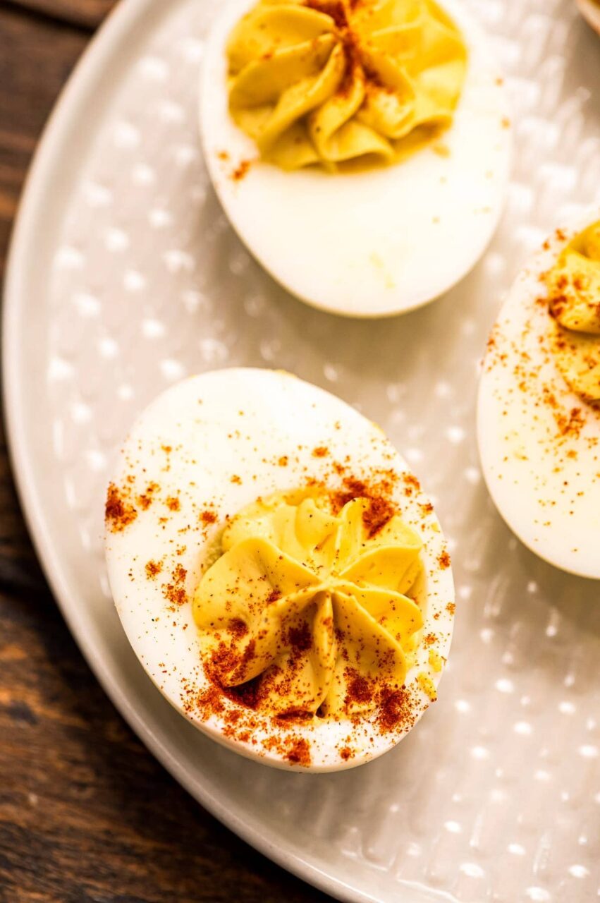 Classic deviled eggs on a white serving platter dusted with paprika and garnished with fresh chives, shot overhead on a rustic wooden picnic table