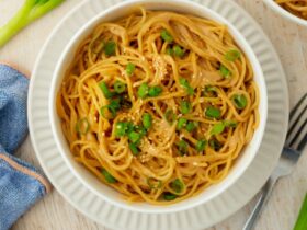 Cold sesame noodles served in a wide bowl topped with sliced cucumbers, shredded carrots, green onions, sesame seeds, and a drizzle of chili oil on a wooden table