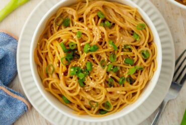Cold sesame noodles served in a wide bowl topped with sliced cucumbers, shredded carrots, green onions, sesame seeds, and a drizzle of chili oil on a wooden table