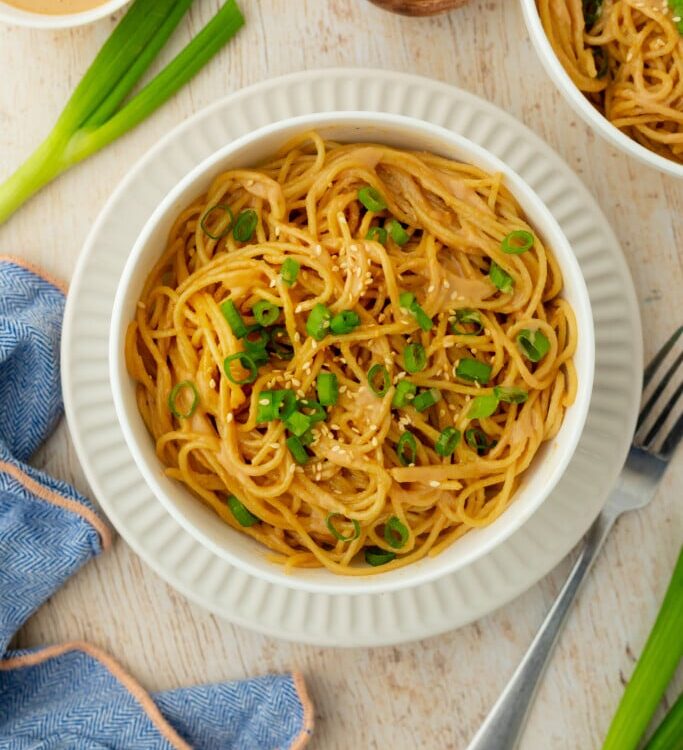 Cold sesame noodles served in a wide bowl topped with sliced cucumbers, shredded carrots, green onions, sesame seeds, and a drizzle of chili oil on a wooden table