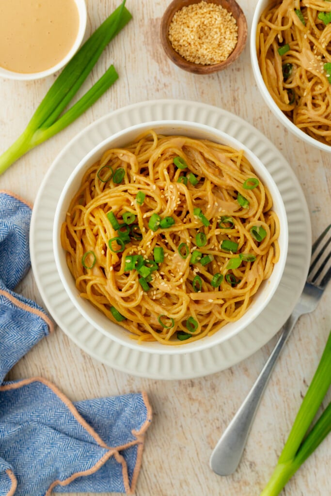 Cold sesame noodles served in a wide bowl topped with sliced cucumbers, shredded carrots, green onions, sesame seeds, and a drizzle of chili oil on a wooden table