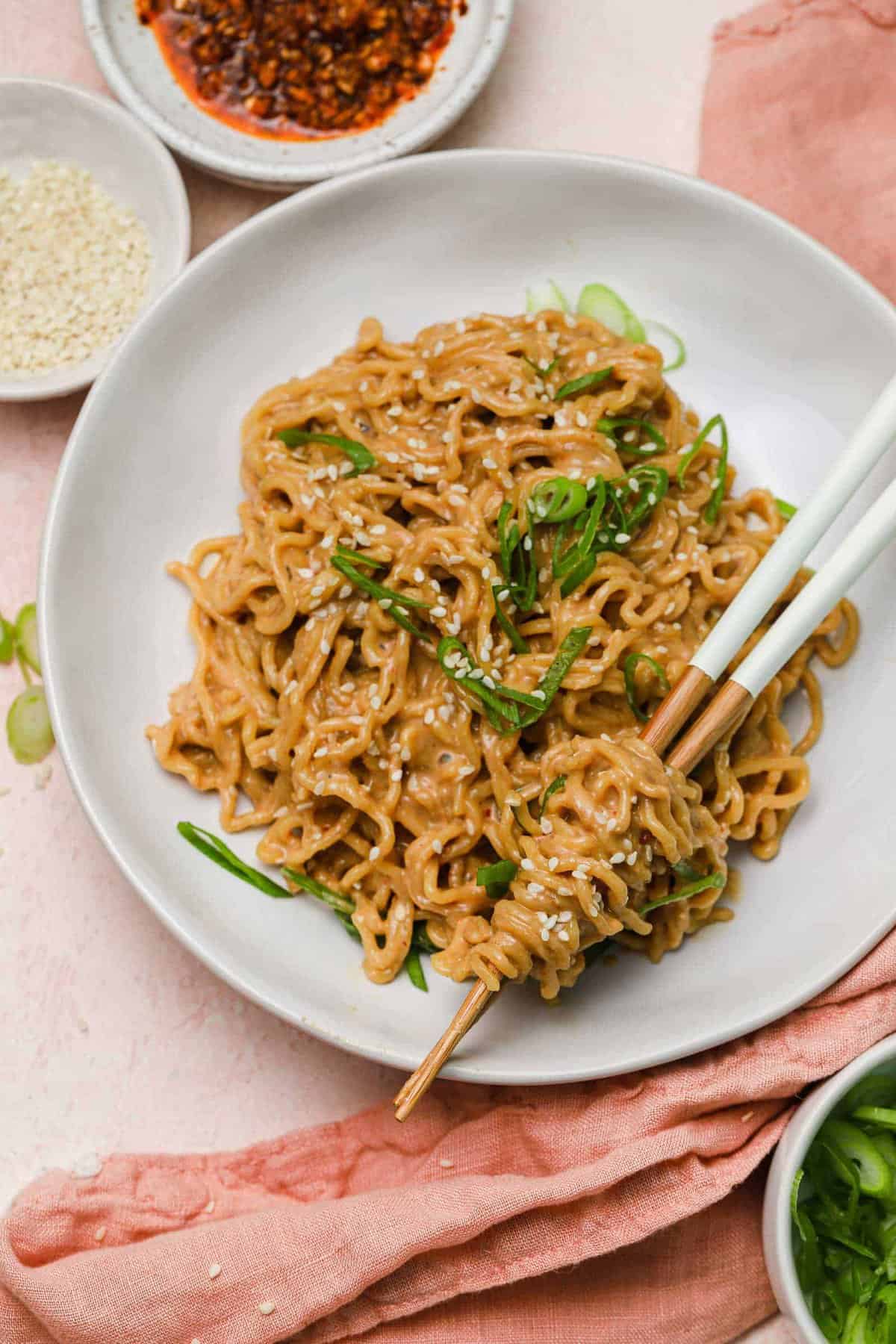Overhead flat lay of cold sesame noodle ingredients including peanut butter, soy sauce, sesame oil, rice vinegar, chili garlic sauce, fresh garlic, ginger, lime, and dry noodles arranged on a white marble surface