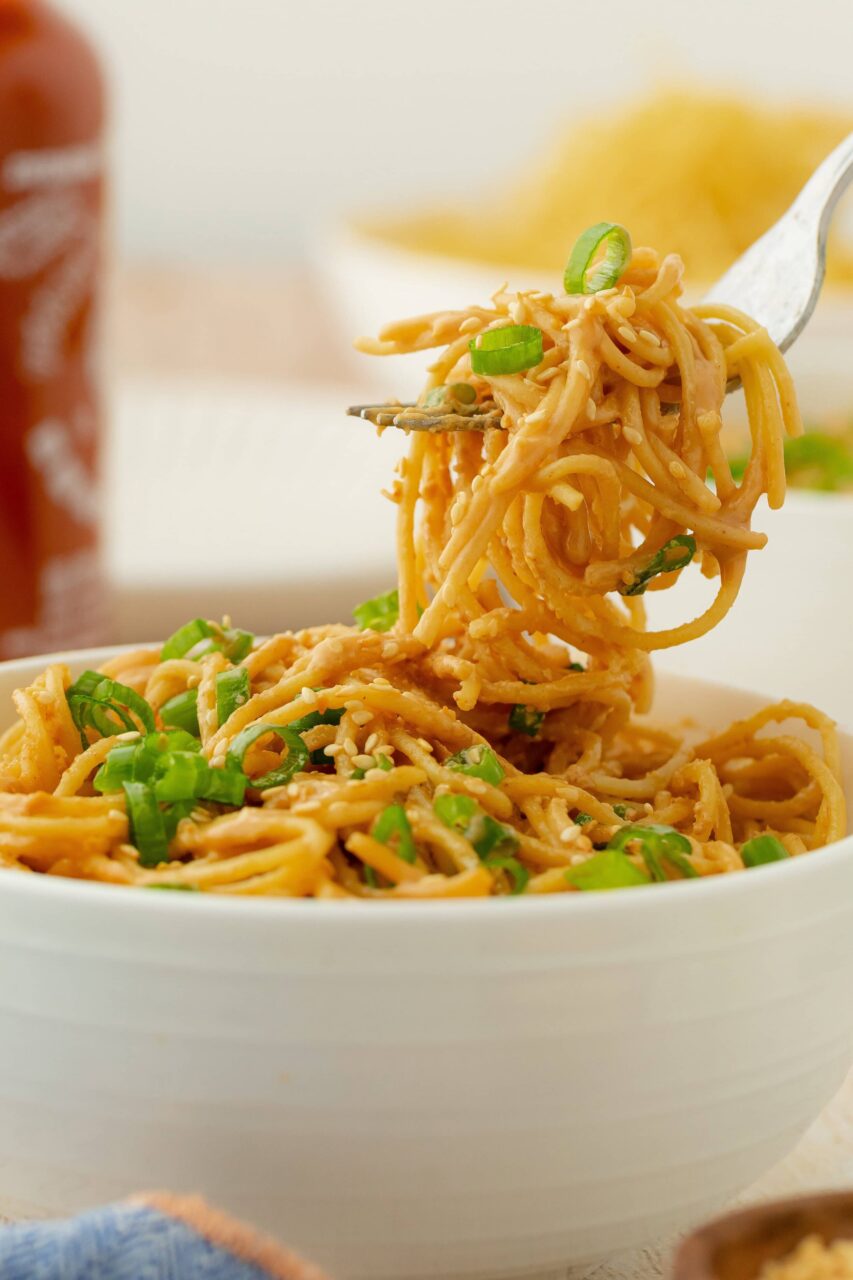 Close-up of cold sesame noodles twirled on a fork with visible peanut sauce coating, topped with sesame seeds, sliced green onions, and a wedge of lime on the side
