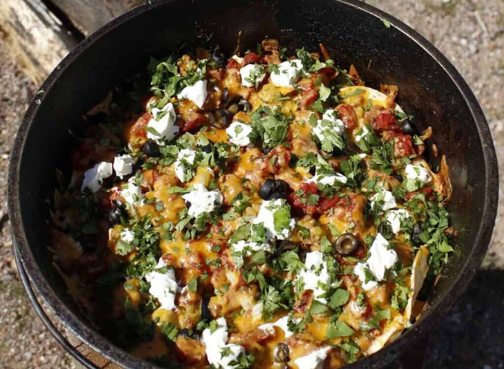 Dutch Oven Campfire Nachos loaded with melted cheese, black beans, salsa, and toppings served straight from a cast iron Dutch oven at a campfire
