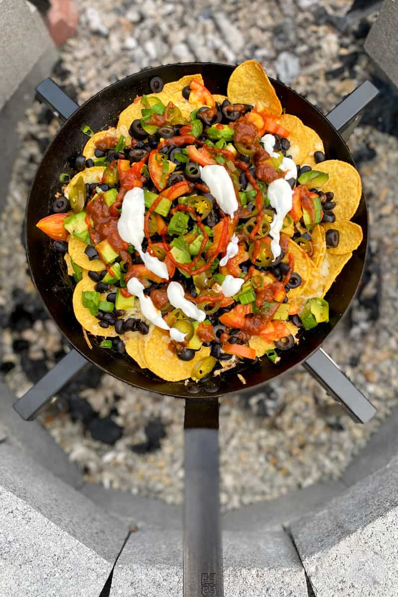 Ingredients for Dutch Oven Campfire Nachos laid out on a picnic table including tortilla chips, shredded cheese, canned black beans, salsa, and toppings