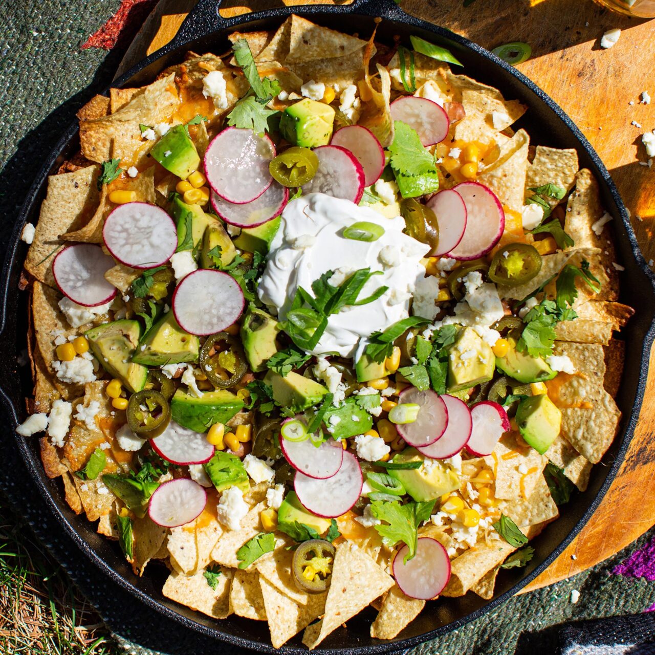 Close-up of Dutch Oven Campfire Nachos topped with sour cream, fresh cilantro, sliced jalapeños, and avocado served on a rustic wooden board