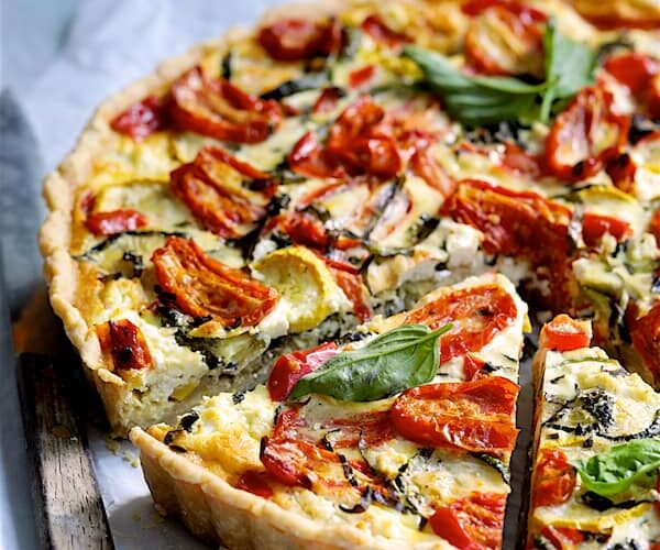 Grilled vegetable and goat cheese tart sliced on a rustic wooden board with fresh herbs and a golden flaky whole wheat crust