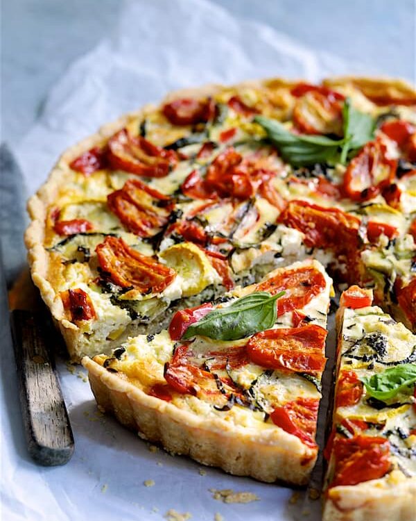 Grilled vegetable and goat cheese tart sliced on a rustic wooden board with fresh herbs and a golden flaky whole wheat crust