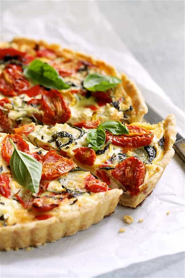 Close-up of a slice of grilled vegetable goat cheese tart on a white plate, showing the flaky whole wheat crust layers, creamy goat cheese filling, and golden charred vegetable topping with fresh thyme