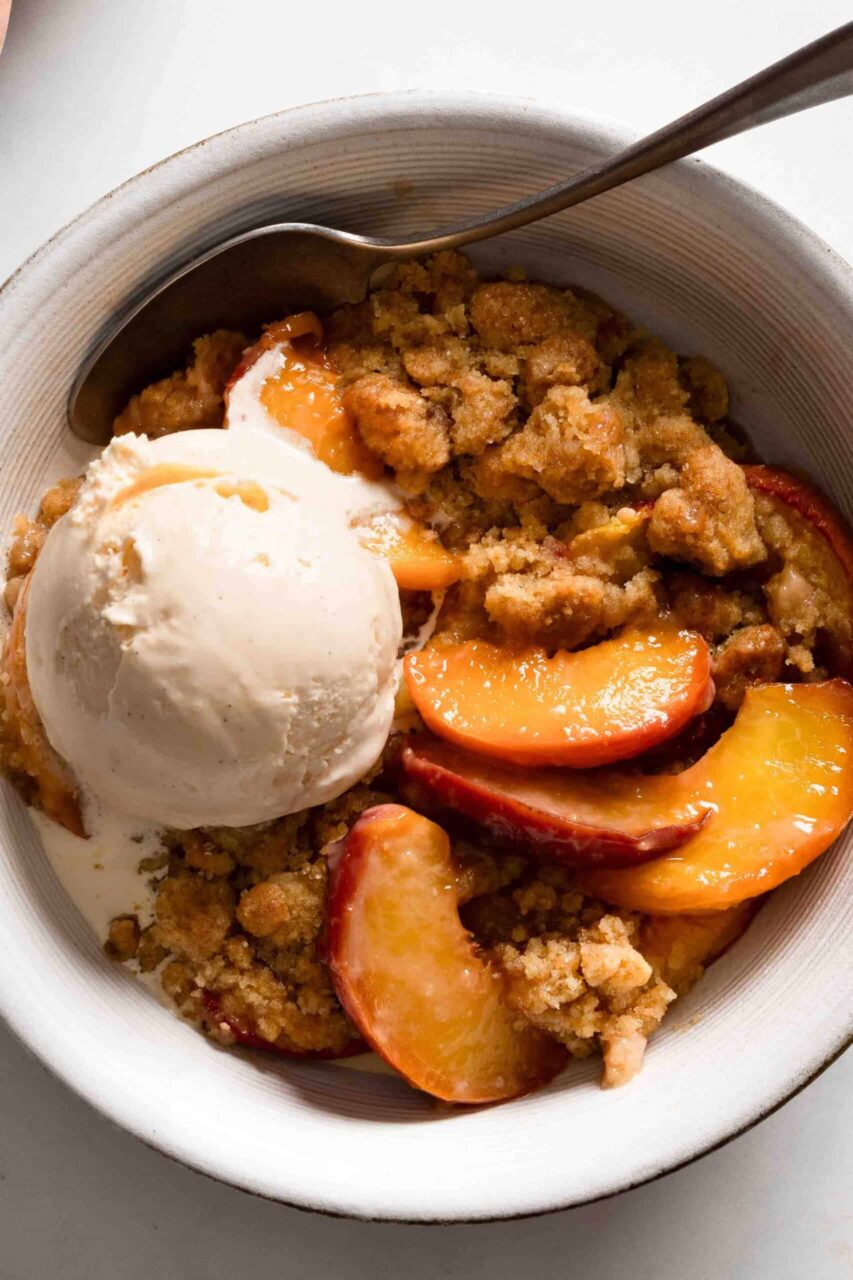 close-up of a scoop of peach cobbler in a white bowl topped with a melting scoop of vanilla ice cream and fresh peach slices