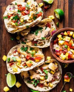 Peach mango salsa in a white bowl garnished with fresh cilantro and lime wedges, served alongside grilled fish tacos on a wooden board
