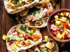 Peach mango salsa in a white bowl garnished with fresh cilantro and lime wedges, served alongside grilled fish tacos on a wooden board