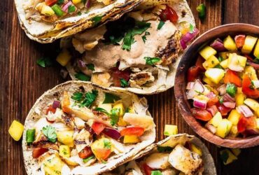 Peach mango salsa in a white bowl garnished with fresh cilantro and lime wedges, served alongside grilled fish tacos on a wooden board