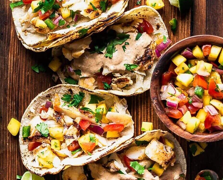 Peach mango salsa in a white bowl garnished with fresh cilantro and lime wedges, served alongside grilled fish tacos on a wooden board