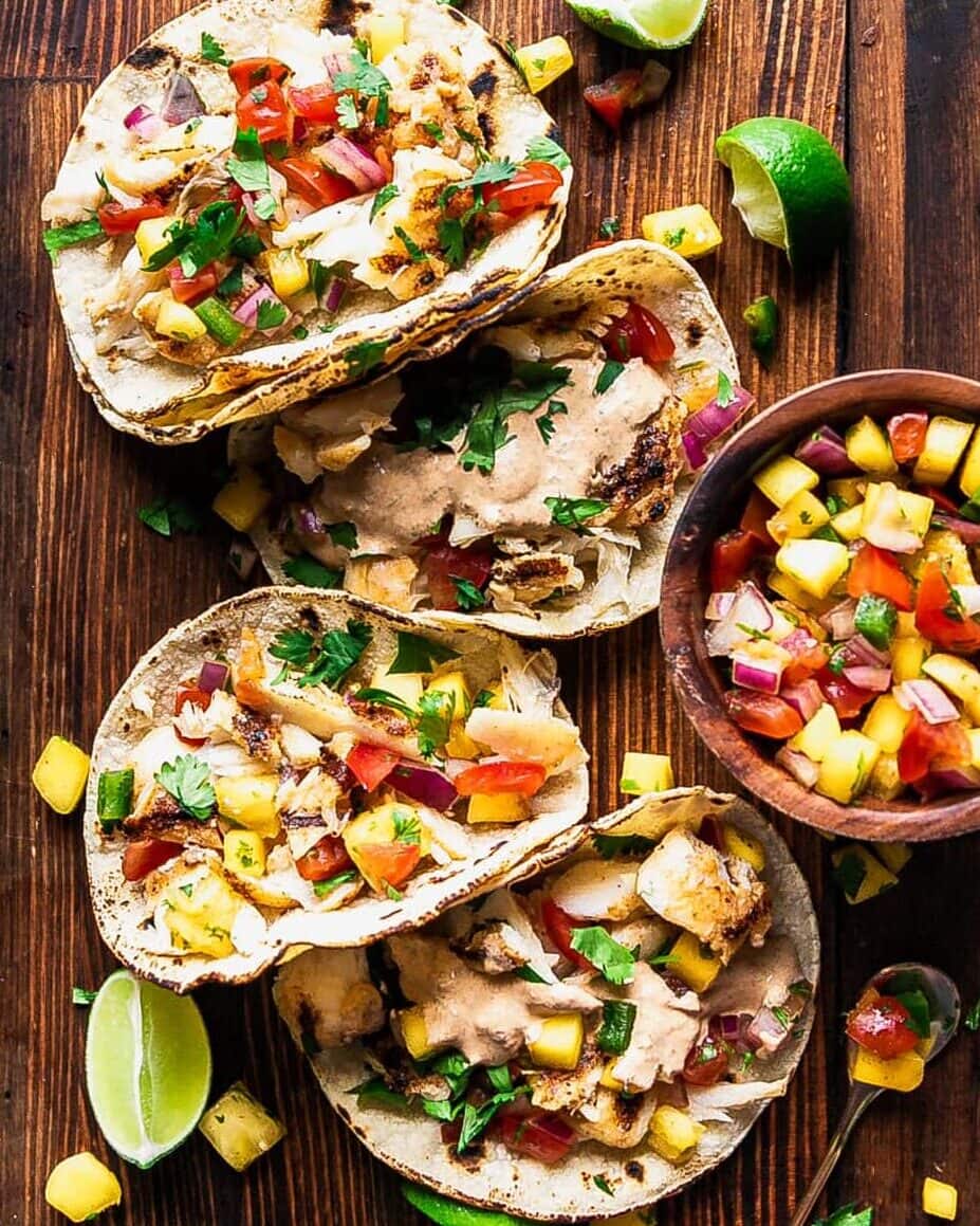 Peach mango salsa in a white bowl garnished with fresh cilantro and lime wedges, served alongside grilled fish tacos on a wooden board