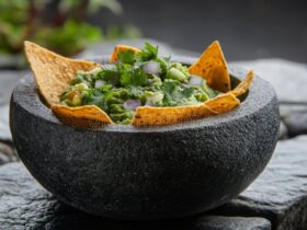 Restaurant style guacamole served in a rustic stone bowl with tortilla chips, garnished with fresh cilantro, diced tomato, and lime wedges on a wooden board