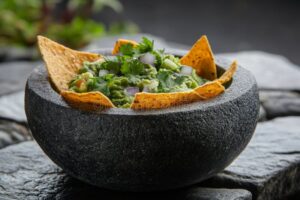 Restaurant style guacamole served in a rustic stone bowl with tortilla chips, garnished with fresh cilantro, diced tomato, and lime wedges on a wooden board