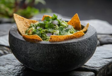 Restaurant style guacamole served in a rustic stone bowl with tortilla chips, garnished with fresh cilantro, diced tomato, and lime wedges on a wooden board