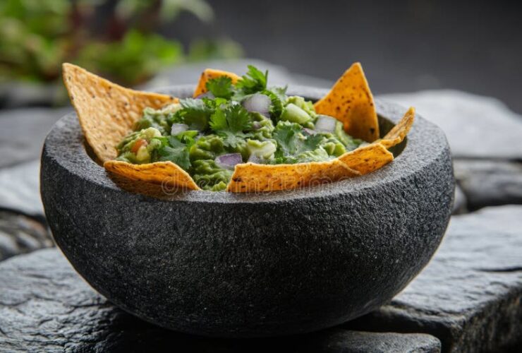 Restaurant style guacamole served in a rustic stone bowl with tortilla chips, garnished with fresh cilantro, diced tomato, and lime wedges on a wooden board