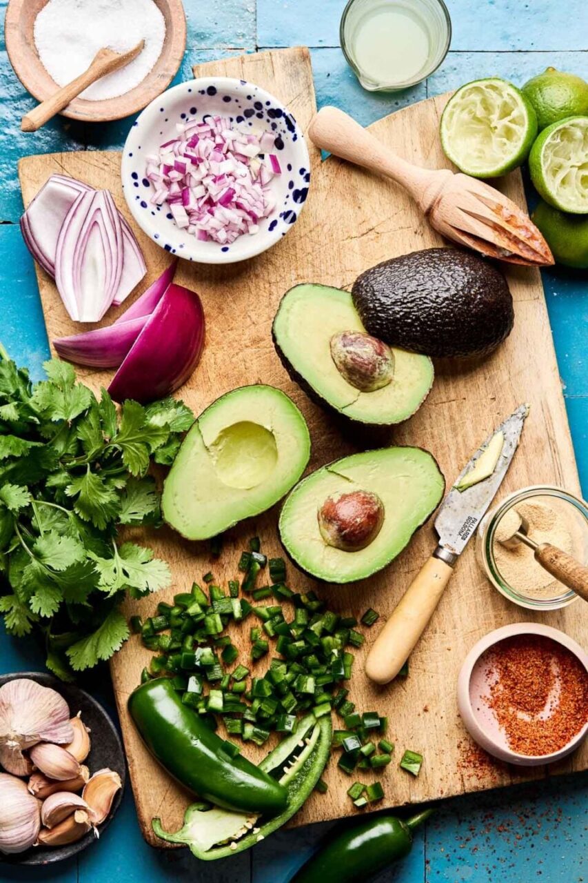 Step-by-step ingredients flat lay for restaurant style guacamole — halved avocados, diced white onion, jalapeño, lime halves, cilantro, and sea salt on a marble surface