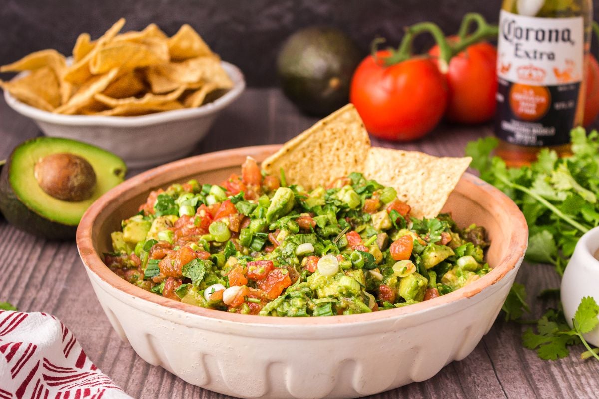Close-up of chunky restaurant style guacamole being scooped with a tortilla chip, showing vivid green avocado chunks, cilantro leaves, and diced red tomato