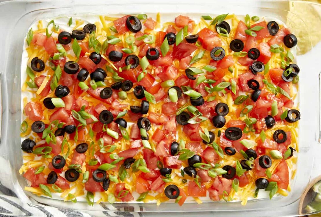 Seven Layer Dip in a large glass dish showing all colorful layers topped with cheese, olives, and tomatoes, served with tortilla chips on a wooden board