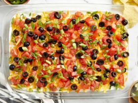 Seven Layer Dip in a large glass dish showing all colorful layers topped with cheese, olives, and tomatoes, served with tortilla chips on a wooden board