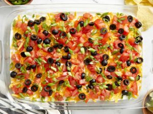 Seven Layer Dip in a large glass dish showing all colorful layers topped with cheese, olives, and tomatoes, served with tortilla chips on a wooden board