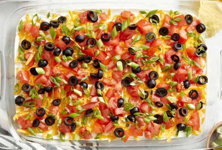Seven Layer Dip in a large glass dish showing all colorful layers topped with cheese, olives, and tomatoes, served with tortilla chips on a wooden board