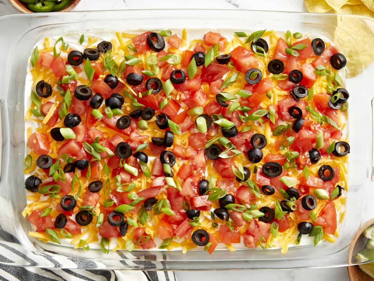 Seven Layer Dip in a large glass dish showing all colorful layers topped with cheese, olives, and tomatoes, served with tortilla chips on a wooden board