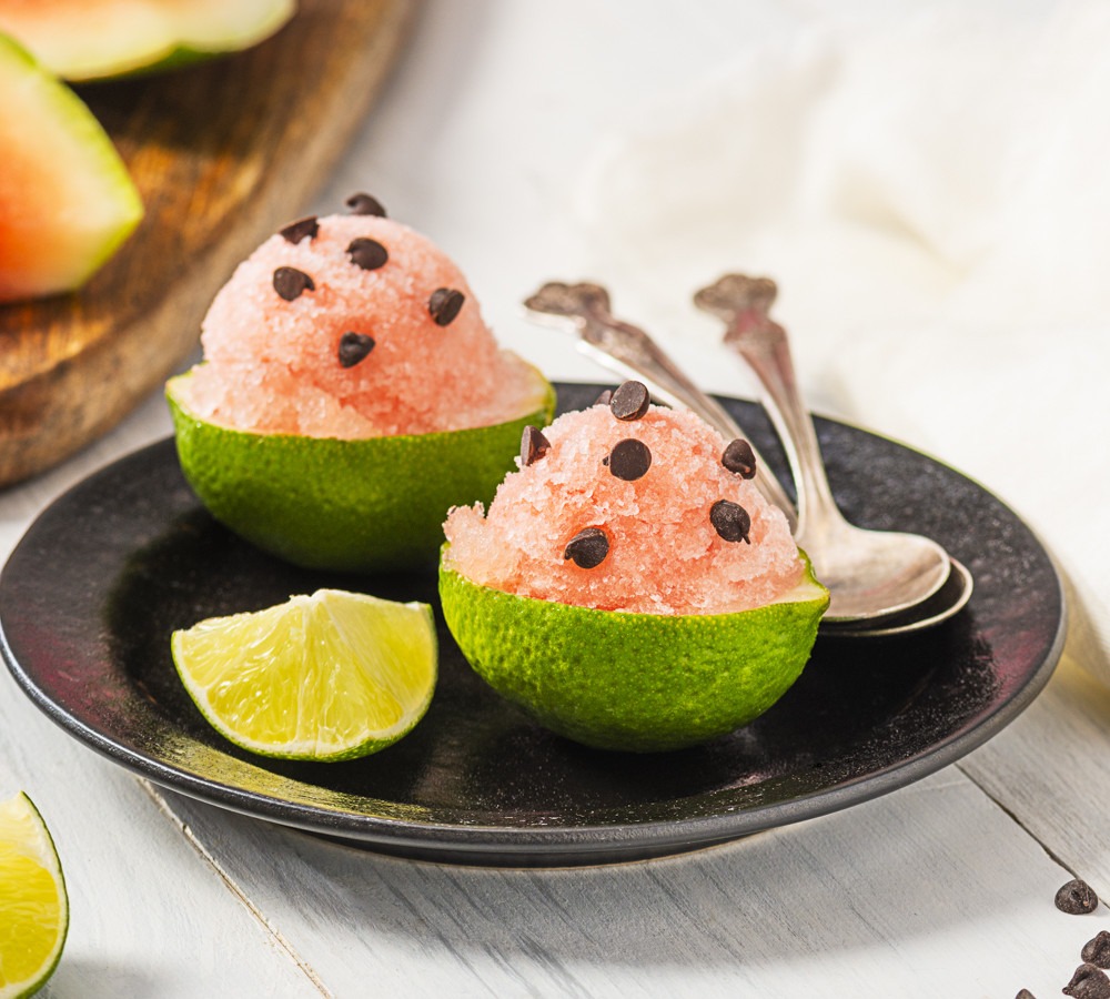 Overhead flat lay of watermelon chunks, halved limes, fresh mint, sugar, and a baking dish with partially frozen granita being scraped with a fork