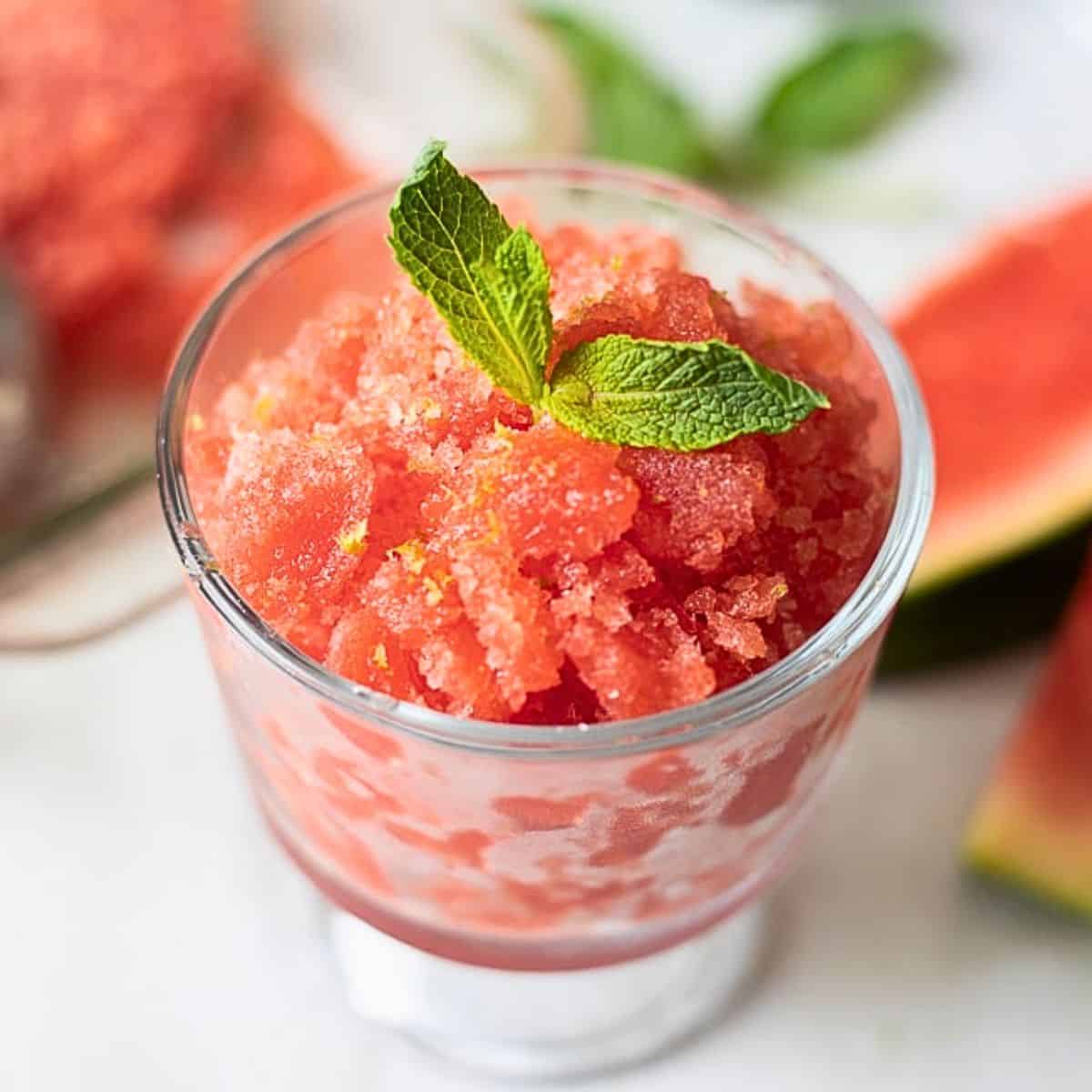 Close-up of watermelon lime granita being scooped into a rustic bowl with visible icy flaky crystals, lime zest scattered on top and mint leaves alongside