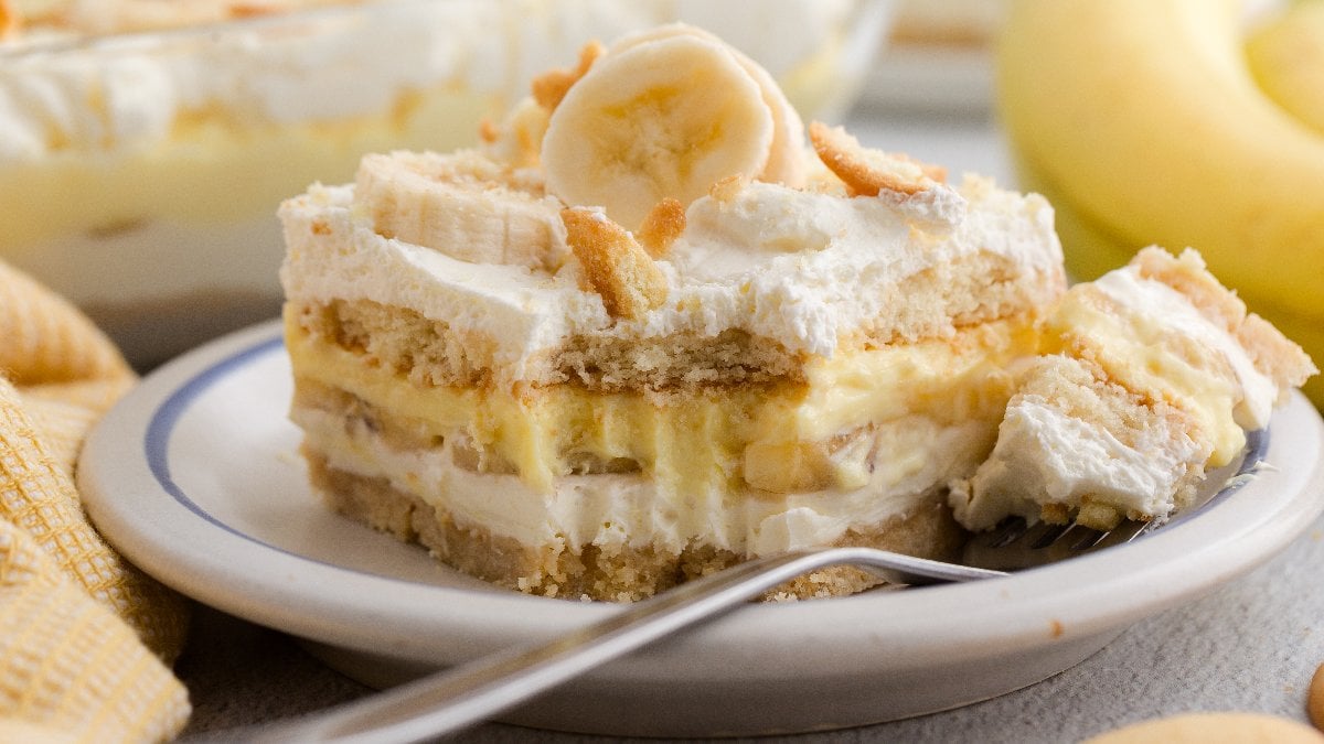 A step-by-step ingredients flat lay showing vanilla wafers, sliced bananas, cream cheese block, pudding mix box, condensed milk can, and whipped topping container arranged on a white marble surface